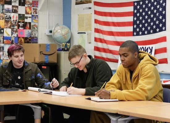 三个学生坐在教室里的一张桌子旁，背景是一面美国国旗. 一个穿黄色连帽衫的学生正在专心写作, 那个戴眼镜的中学生也在写字, 第三个染了头发的学生正在抬头看.