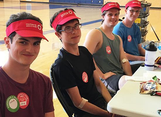 四个肤色浅的年轻人, 戴着红色的美国红十字会面罩, 坐在体育馆的桌子旁. 他们穿着便服，贴着参加献血活动的贴纸.